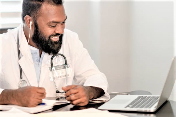 Picture of a black doctor smiling at a computer at a table. 